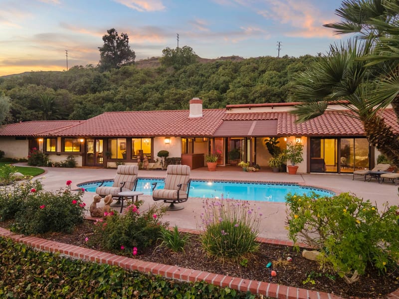 Spanish tile roof ranch home glowing at twilight behind a sparkling pool and rose garden