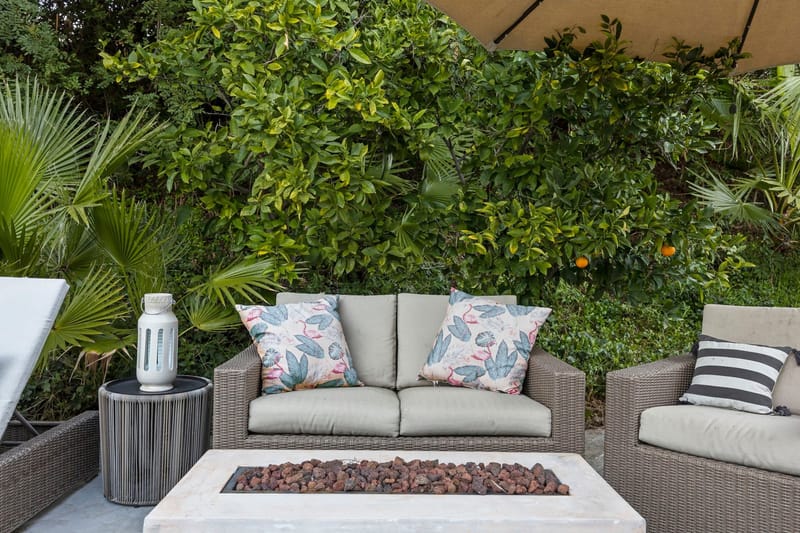 Outdoor wicker lounge seating with tropical pillows around a stone gas fire pit beside citrus trees