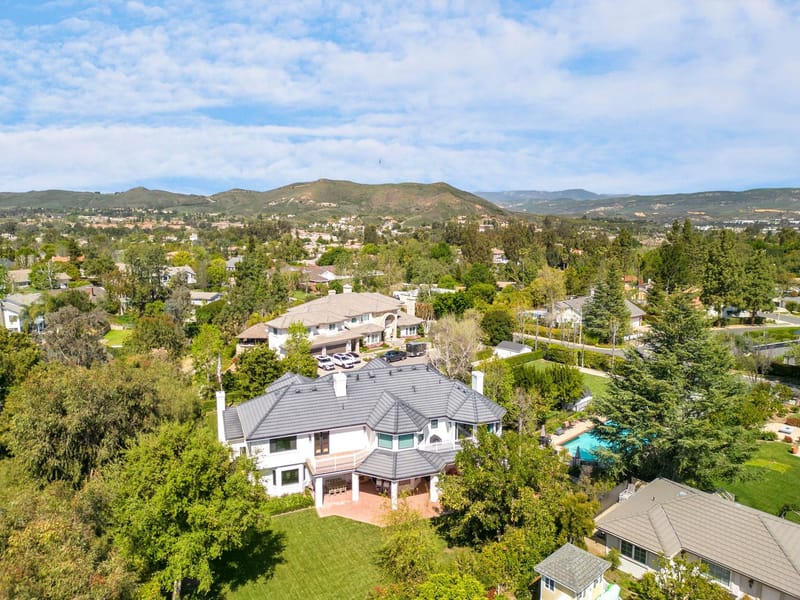 Aerial view of a large white estate with gray tile roof in a green hillside neighborhood