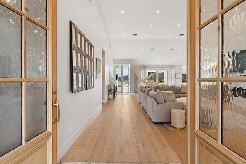 Entry view through glass doors into a bright living room with wide plank oak floors and hillside views