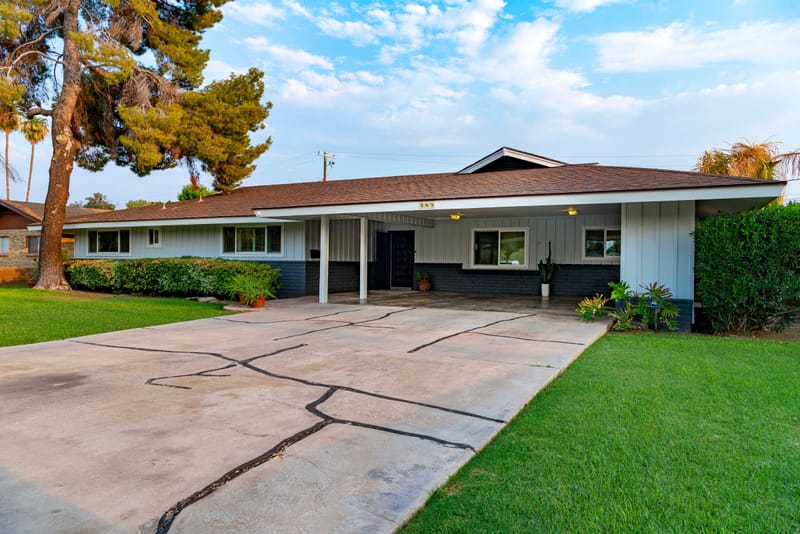 Single story midcentury ranch home with carport, wide concrete driveway and green lawn under blue sky