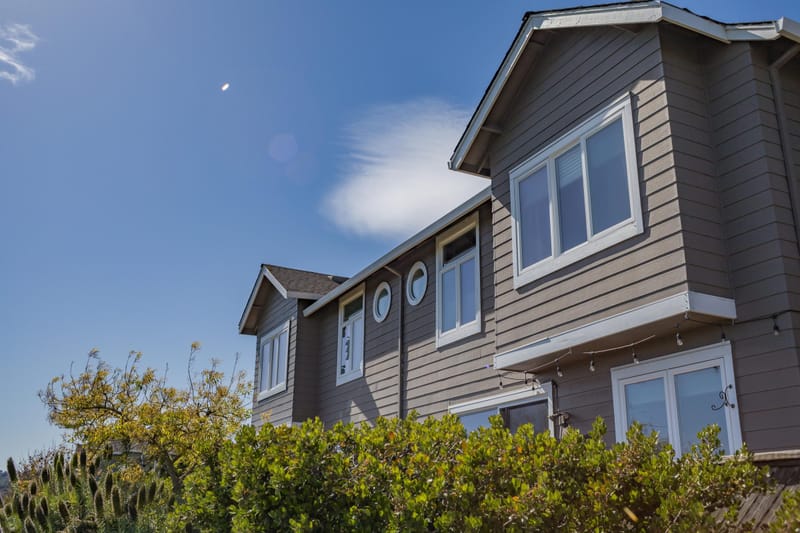 Gray shingled two story coastal home exterior with white trimmed windows under a clear blue sky