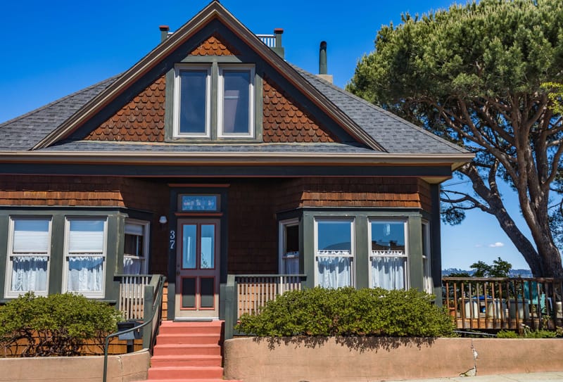 Shingled craftsman cottage with green trim, red steps and a cypress tree under deep blue sky