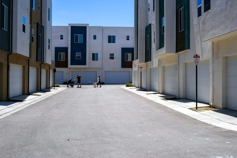 Modern townhomes line a sunlit alley of garages under clear blue California sky as a family walks past.de