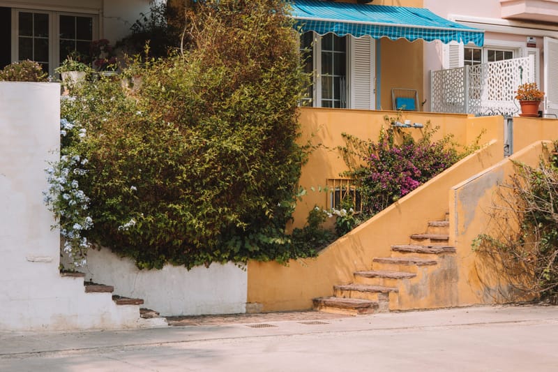 Sunlit Mediterranean style home with yellow stucco walls, stone steps and bougainvillea blooms along a quiet street.?