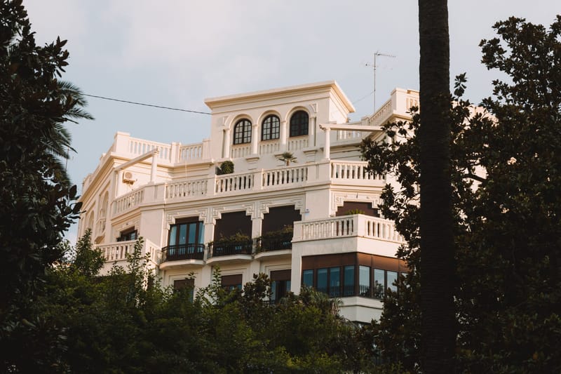 White Mediterranean style estate with arched windows and balustrades framed by lush trees at golden hour.c