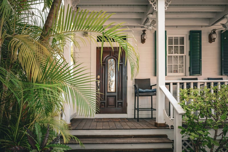 Wooden front door of a white cottage framed by palm fronds and a shaded porch with green shutters.-->