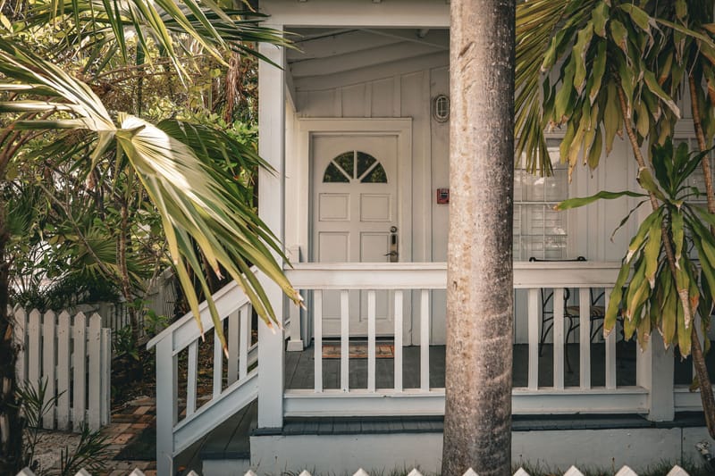White cottage porch with railing and arched door framed by palm fronds and picket fence at dusk.ec
