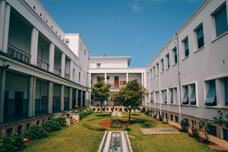 Sunlit white institutional building framing a green courtyard with citrus trees and a narrow water channel