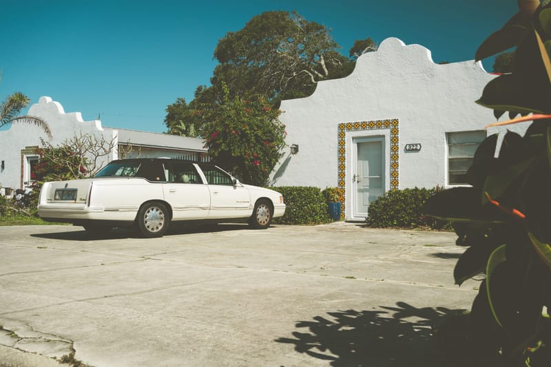 White Spanish style bungalow with scalloped parapet, tile trimmed doorway and vintage sedan in the driveway under blue sky.$