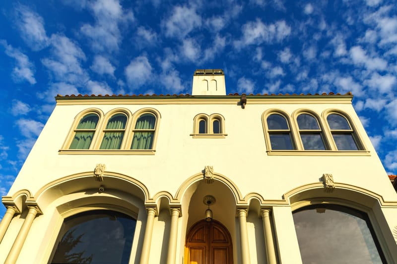 Spanish revival stucco home facade with arched windows and tile roof under a bright blue sky.md