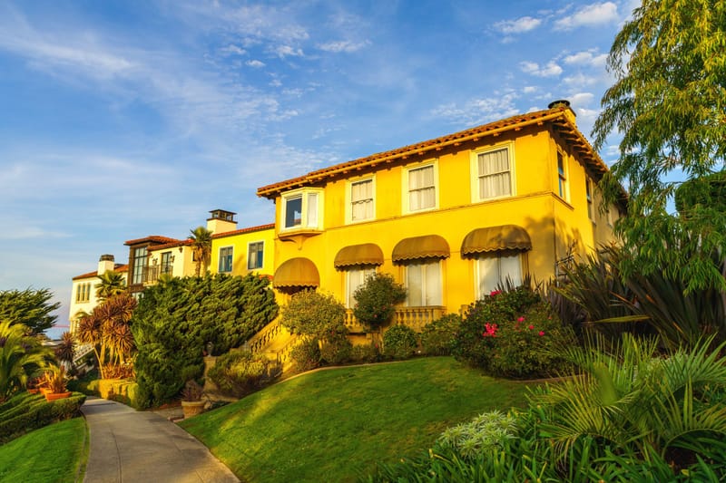 Golden hour light on a yellow Mediterranean style hillside estate with tile roof, awnings and lush gardens.์
