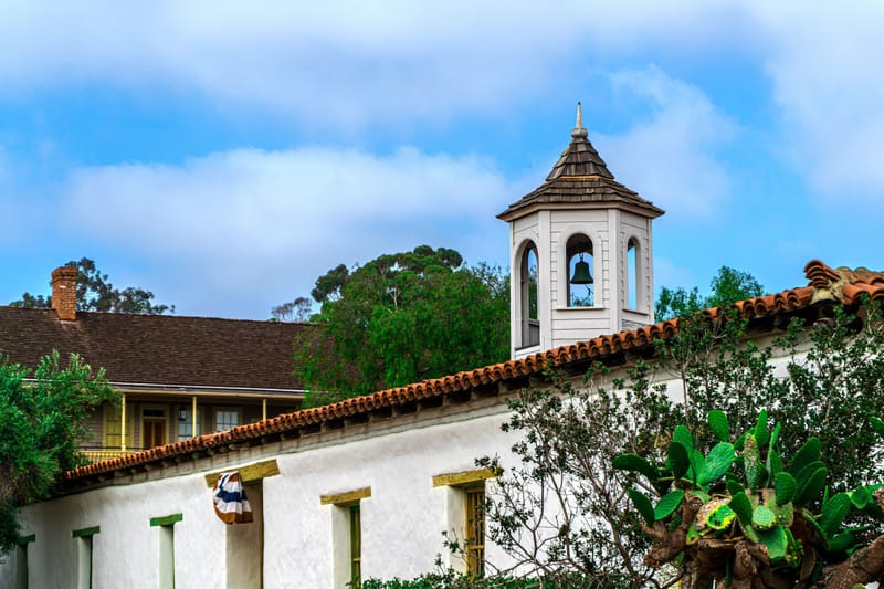 Spanish colonial adobe building with red tile roof, white bell tower and cactus under blue California sky','