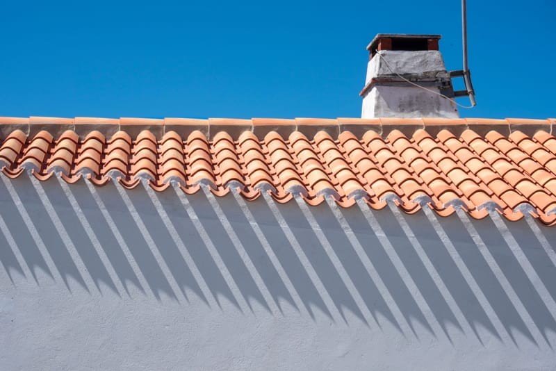 Terracotta clay roof tiles casting sharp shadows on a white stucco wall under deep blue sky.;s chimney above.)
