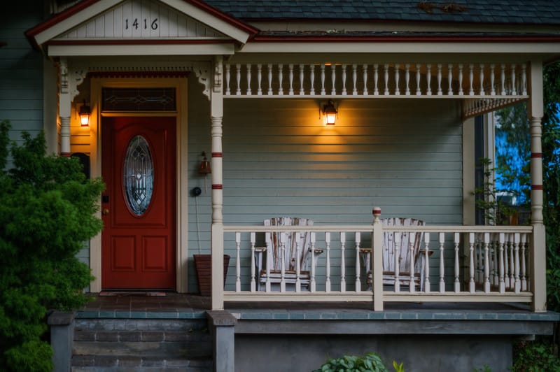 Victorian craftsman home porch at dusk with red front door, warm lantern light and white railing chairs.Victorian craftsman porch at dusk with red door and warm lanterns