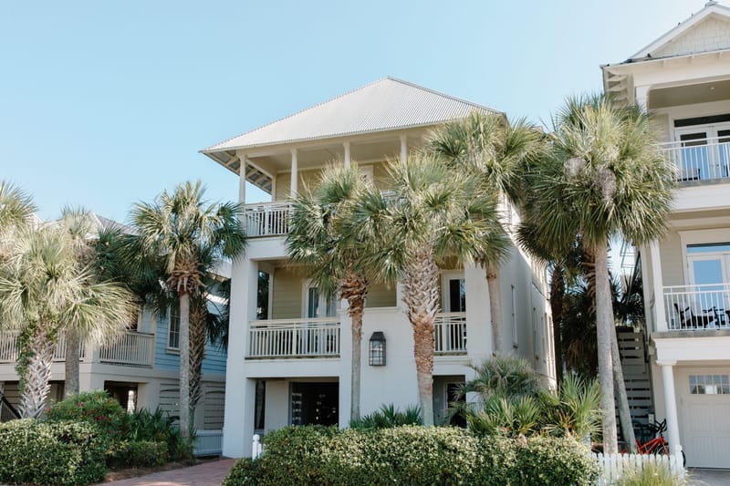 Three story coastal beach houses with white railings framed by tall palm trees under a clear blue sky