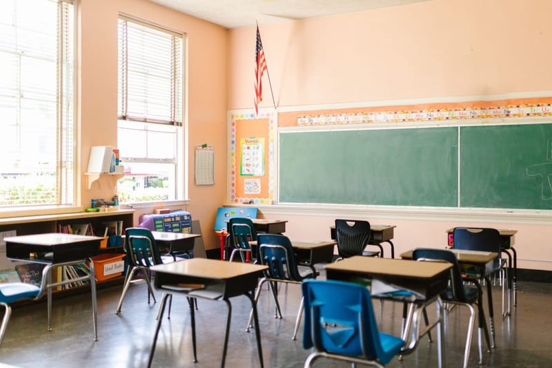 Empty elementary classroom with wooden desks, green chalkboard and sunlight through tall windows.-